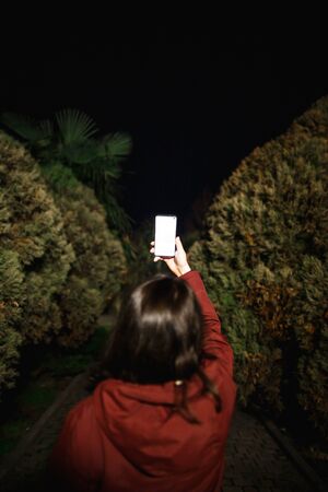 Woman with phone at night in the park is a view from behind. Theres a dark alley ahead