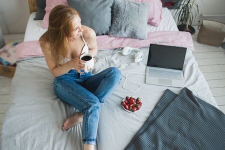 Blonde sexy woman with a cup of tea or coffee and strawberries having fun in bed. She wearing white bra and casual jeans. Happy morningの写真素材