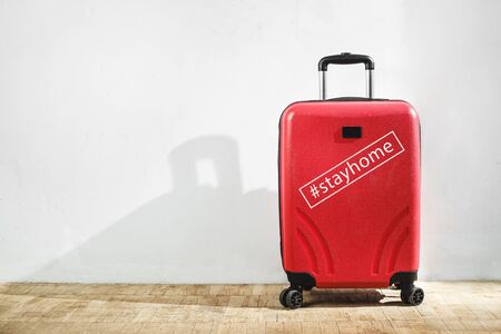 Red suitcase with inscription tag stayhome against the background of a light wall in the room. Stay home, travel concept. Copy spaceの写真素材