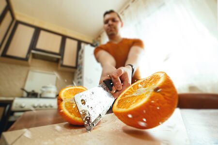 Man cutting juicy orange in half with splashes in kitchen. Wide angle closeup. Creative lockdown