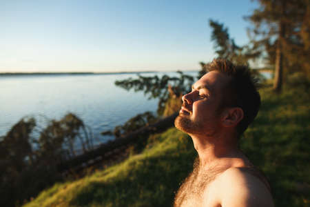 Handsome bearded caucasian man with closed eyes standing on cliff at beautiful sunset. In background is river and taiga forest.の写真素材