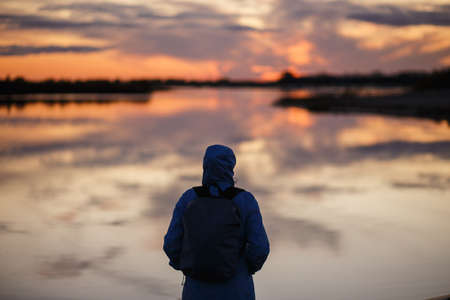 Silhouette of a young woman in autumn jacket with hood standing on the river or lake shore at...