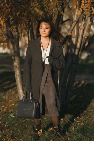 Stylish caucasian girl in the fur coat with suitcase at beautiful sunny autumn alleyの写真素材