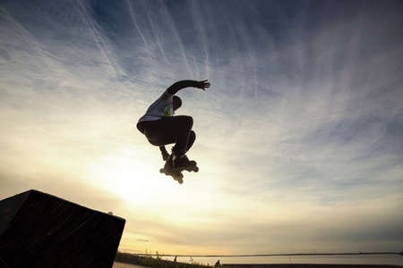 Unrecognizable roller on the extreme roller skates performing acrobatic jump at sunny sky - Guy at extreme sport competition on sunny afternoon - Alternative lifestyle concept on warm sunshine colors. Warm toned image.の写真素材