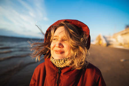 Beautiful girl with wild hair in hood looking to the sunset on the sea coast. Sunny winter day on...