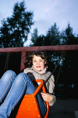 Beautiful young girl energetically and with pleasure swings on the swing at dusk
