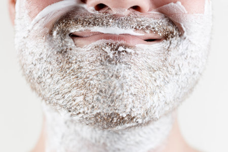 Closeup of male face. Men's beauty. Smiling Young man is applying moisturizing and anti-aging cream on his face against white backgroundの写真素材
