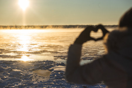 Woman making heart with hands outdoors in a frosty sunset, closeup. Focus on ice floes. High quality...