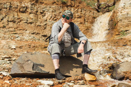 A man in shorts and a hat is leisurely sitting on a bedrock outcrop near a majestic waterfall in the...