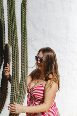 A stylish woman poses in a chic striped dress beside a tall cactus, framed by a bright white wallの写真素材