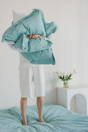 A person is currently standing on a bed while holding a fluffy pillow high above their head,...
