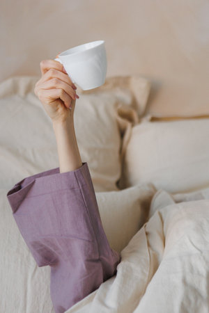 A person is comfortably laying in bed while holding a warm cup of coffee, enjoying a relaxed moment...