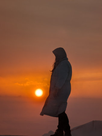 A serene silhouette of a person in a hooded jacket, gazing at a vibrant sunset over the horizon, evoking feelings of contemplation and tranquility.の写真素材