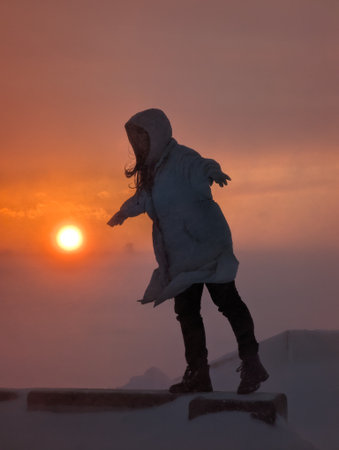 A girl wearing a warm coat joyfully balances on a snowy edge against a stunning sunset backdrop, capturing the essence of adventure and winter beauty.の写真素材