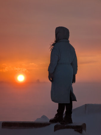 A silhouetted figure stands on a snow-covered surface, gazing at a breathtaking sunrise, evoking a sense of tranquility and wonder in a winter landscape.の写真素材
