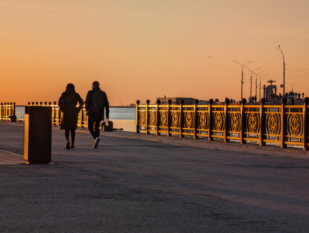 A couple walks hand-in-hand along a picturesque waterfront at sunset, enjoying a peaceful moment together against a vibrant sky.の写真素材