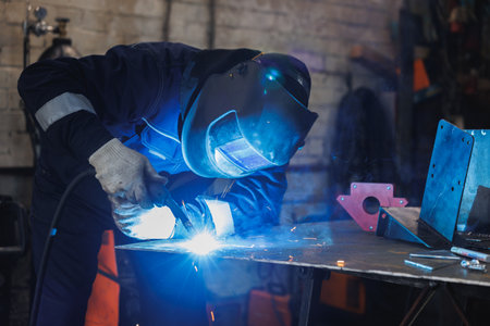 In a bustling factory, a skilled man is carefully welding a piece of metal, ensuring safety by...