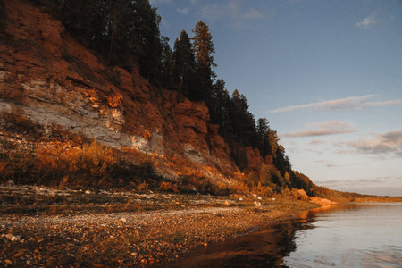 A stunningly tranquil riverbank scene showcases a dramatic cliffside and vibrant natural hues during sunset hoursの写真素材