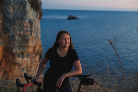 Cyclist Resting With Road Bike By Cliff Overlooking Calm Sea At Twilight, Contemplative Expression, Softの写真素材