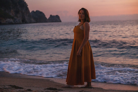 Woman On Sandy Beach At Sunset, Standing Barefoot In Flowing Rust Dress, Waves Lapping, Dramatic...