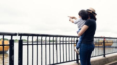 Asian mother and her toddler girl boy pointing out to the big boats in the river while answer the question from her daughter, learning and explore the environment together, happy family outing time.の写真素材