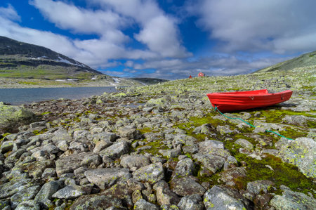 a red fishing boat on the shore of a lake from melted snow on the snow road in Norway with red wooden houses in the background during a summer dayの写真素材