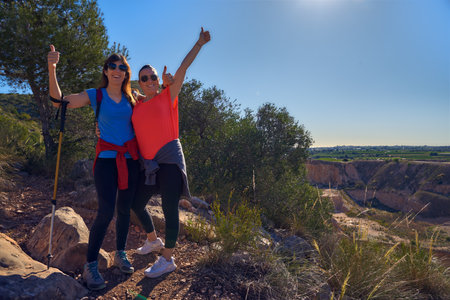 two young women hiking and happy holding together on a trail in the mountain rising their arms in a summer dayの写真素材