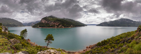 panoramic view of a water reservoir with cloudy sky and surrounded by mountainsの写真素材
