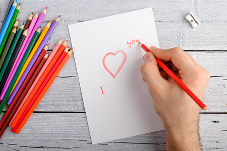 drawing with colored pencils postcard for Valentine's day right-hand on white paper on white wooden background, copy space for text, top view.の写真素材