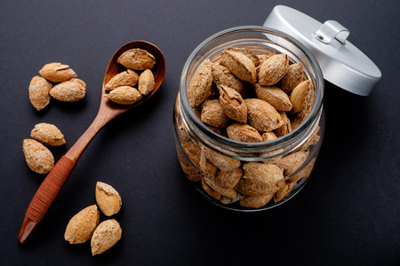 salted almonds in shell in glass jar and brown wooden spoon on black slate backgroundの写真素材