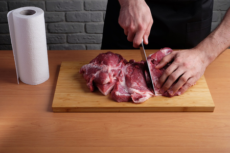 man in the black apron cuts of pork or beef on a cutting wooden board in the kitchenの写真素材
