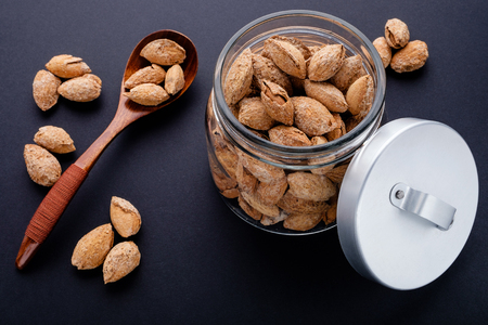 salted almonds in shell in glass jar and brown wooden spoon on black slate backgroundの写真素材