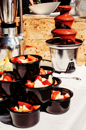 chocolate fountain and slice strawberries banana in black cups on a white table, delicious sweets dessert, street food, outdoorの写真素材