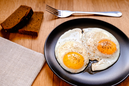 two fried egg with black pepper on black plate on wooden tableの写真素材