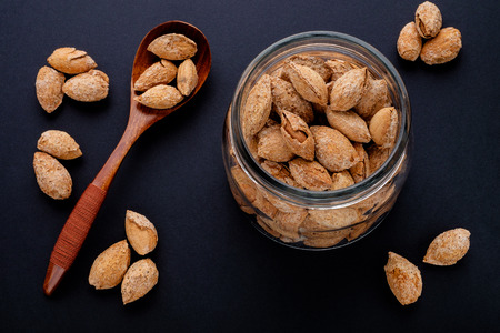 salted almonds in shell in glass jar and brown wooden spoon on black slate backgroundの写真素材