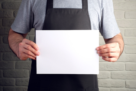 man in black apron holds white sheet of paper in his hands, on background of brick wall mock upの写真素材