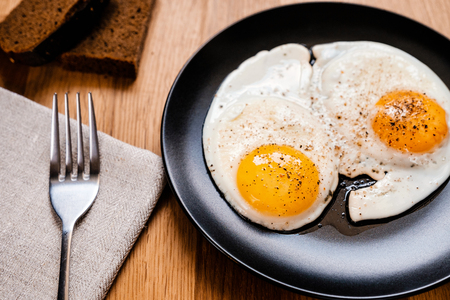two fried egg with black pepper on black plate on wooden tableの写真素材
