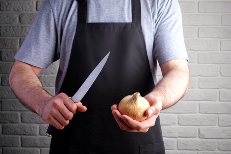 man in black apron holding a knife and onions in his hands, on background of brick wallの写真素材