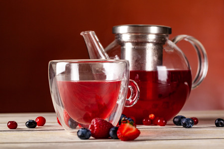 Mint tea with forest berries in a cup of double glass and a glass teapot on a wooden table, on a red...