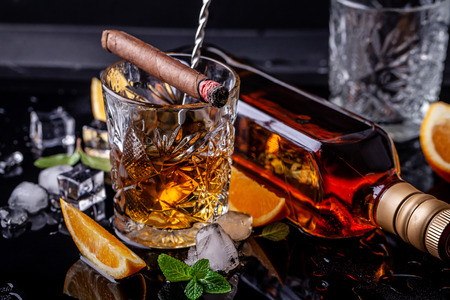 concept of alcoholism, loneliness, dependence. A bottle of whiskey, a glass of whiskey and with ice, a cigar with smoke on a black table, on a black background. Elite drink for masculine relaxationの写真素材