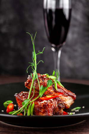 The concept of American cuisine. Roasted pork ribs, baked and glazed in barbecue sauce. Serving dishes in the restaurant on a black plate with sesame and micro green. Next to a glass of red wine.の写真素材