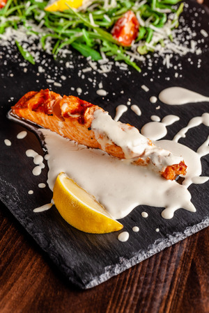 The concept of Italian cuisine. Salmon fillet with arugula, cherry tomatoes and parmesan cheese in creamy lemon sauce. A glass of white wine on the table. Dishes in the restaurant.の写真素材