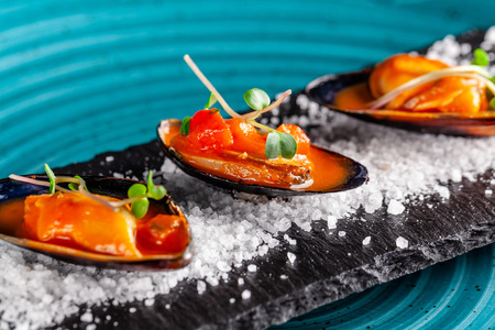 European cuisine. Marinated mussels in tomato sauce with rosemary, garlic, chili. Serving dishes in the restaurant on a plate with coarse sea salt on a black background. Copy spaceの写真素材