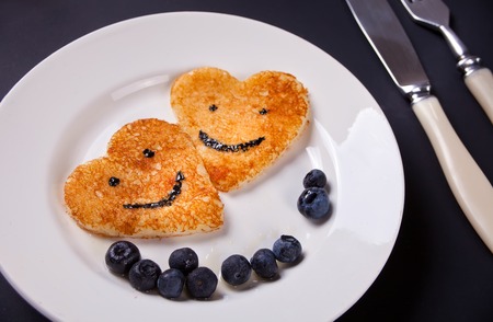 plate of two pancakes with smiles in the shape of heart with berries on white tableの写真素材