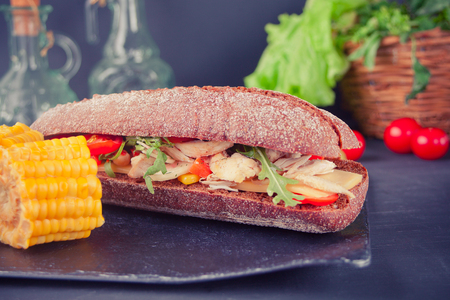 Baguette sandwich with turkey breast, cheese, lettuce, arugula, tomatoes and onion on a black plate on the tableの写真素材