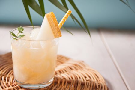 Glass with melon cocktail with straw on a white table with palm leaf in tropical themeの写真素材