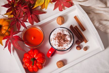 Red mug of hot creamy cocoa with froth on the wooden tray with cookies, autumn leaves, candle and pumpkin on the background. Top view.の写真素材