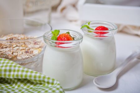 Two portions natural homemade yogurt in a glass jar with fresh strawberry and muesli nearby.の写真素材