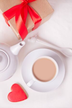 Cup of coffee and a heart shaped red cookie with gift box and teapot on the white table. Top view.の写真素材