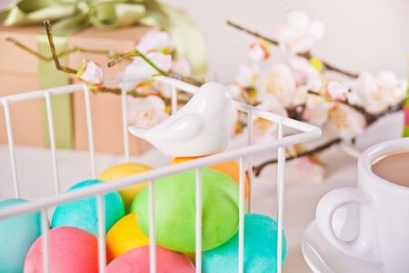 Colorful eggs for Easter day in a basket. Cherry blossom branch, cup of tea and gift box on the...